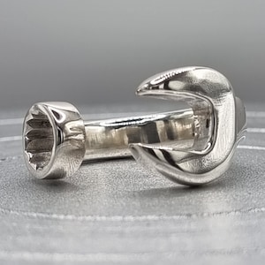Puede incluir: Un anillo de plata con forma de llave inglesa. El anillo está abierto por un lado y tiene un extremo redondeado por el otro.