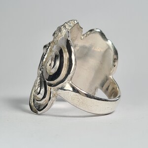 May include: A silver ring with a detailed, swirling design. The ring is wide and has a smooth, flat surface on the top.