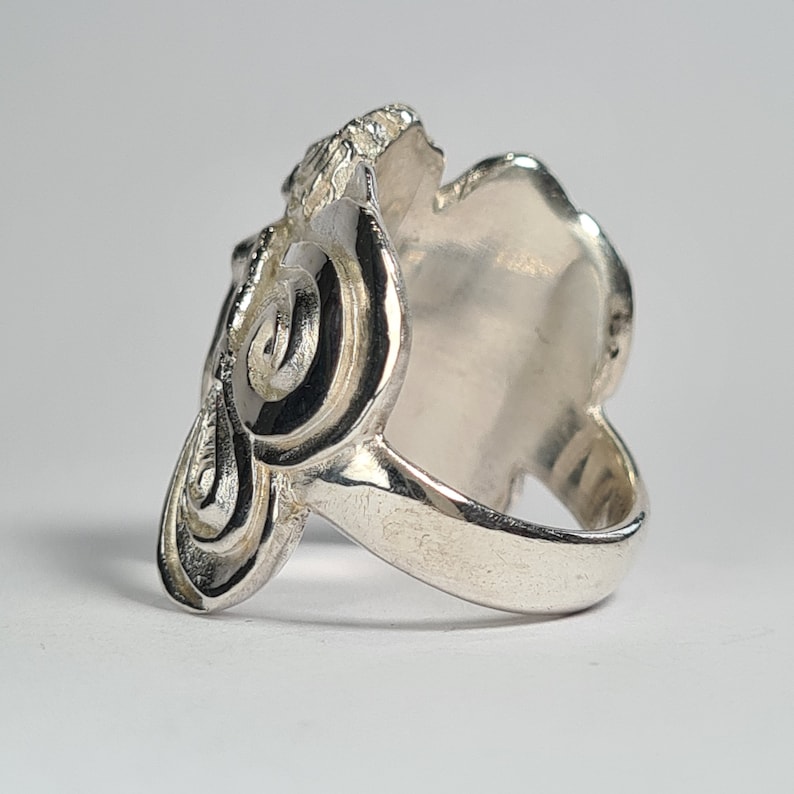 May include: A silver ring with a detailed, swirling design. The ring is wide and has a smooth, flat surface on the top.