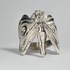 May include: A silver ring with a detailed design of a fairy with wings spread out. The fairy is standing on a ring band.