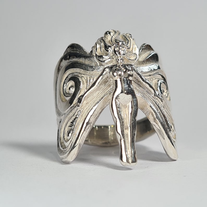 May include: A silver ring with a detailed design of a fairy with wings spread out. The fairy is standing on a ring band.