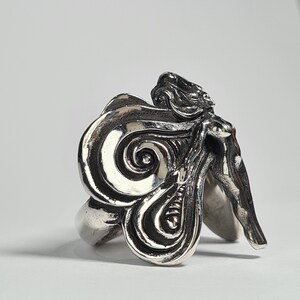 May include: A silver ring with a detailed design of a woman's figure with flowing hair and a swirling pattern.