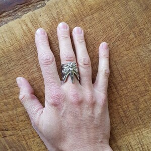 May include: A silver ring with a detailed design of a winged creature with a human-like face. The ring is worn on the middle finger of a hand.
