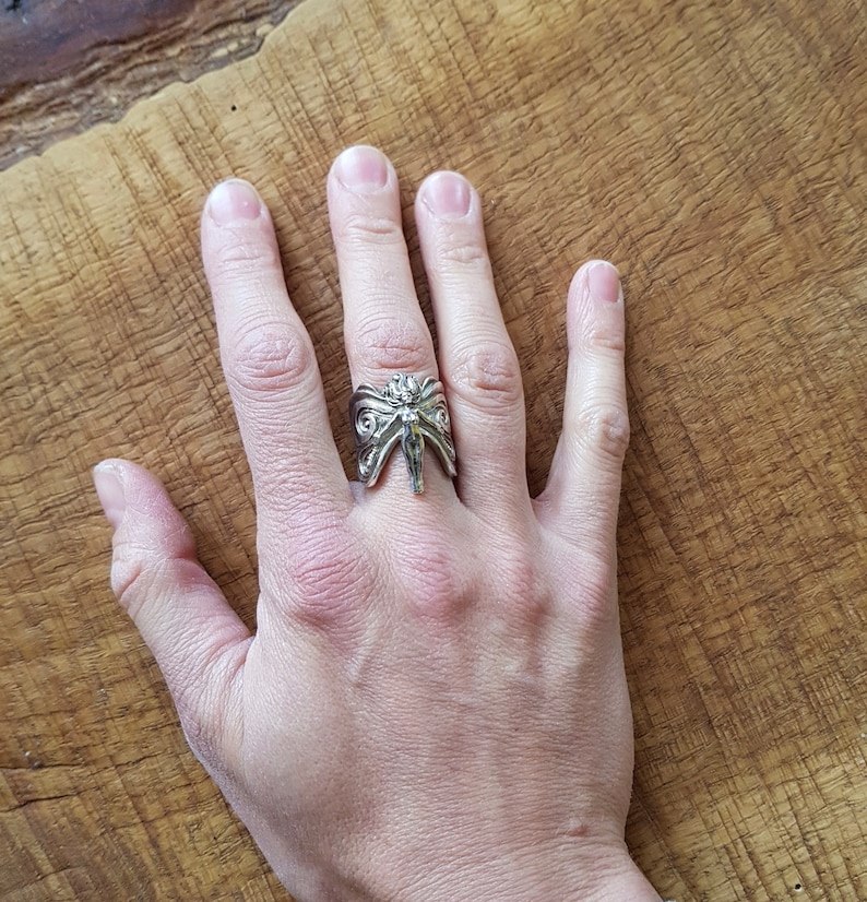 May include: A silver ring with a detailed design of a winged creature with a human-like face. The ring is worn on the middle finger of a hand.