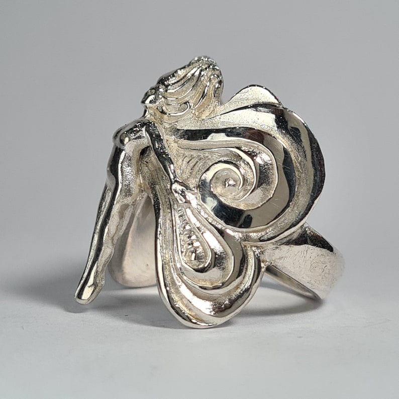 May include: A silver ring featuring a detailed sculpture of a woman with flowing hair and wings. The ring is made of a smooth, polished metal and has a delicate design.