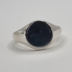 Könnte beinhalten: Ein silberner Siegelring mit einem dunkelblauen, oval geformten Stein. Der Ring hat eine polierte, reflektierende Oberfläche und ein klassisches, minimalistisches Design. Der Stein scheint eine strukturierte Oberfläche zu haben.