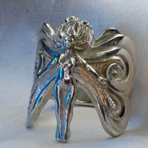 May include: A silver ring with a detailed design of a woman with wings. The woman is standing with her arms outstretched and her wings are spread out behind her.