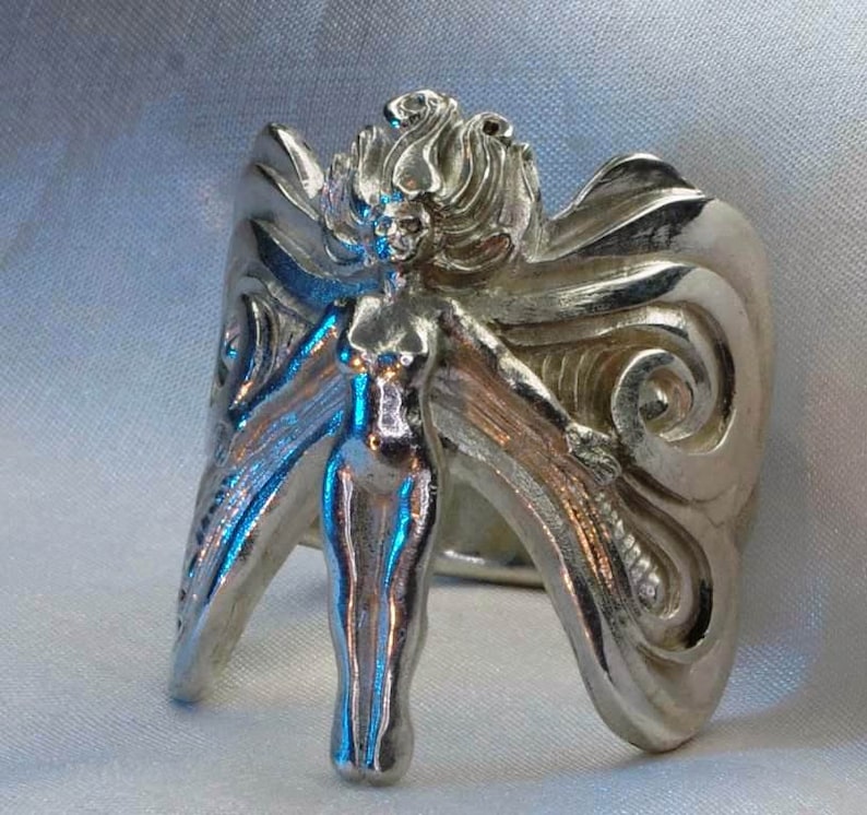 May include: A silver ring with a detailed design of a woman with wings. The woman is standing with her arms outstretched and her wings are spread out behind her.