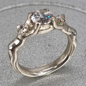 以下が含まれることがあります： 2匹の猫の姿がクリアな宝石を支えるシルバーリング。リングは、中央の石を支えているように見える猫が特徴の、詳細な彫刻デザインです。バンドは滑らかで丸みを帯びています。