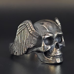 May include: A silver skull ring with wings on the side. The skull is detailed with a prominent nose and eye sockets.