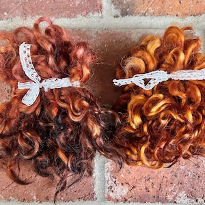 May include: Two bundles of curly, reddish-brown hair, each tied with a white lace ribbon. The hair has varying shades of red, orange, and brown, creating a natural, textured appearance. The background is a brick wall.