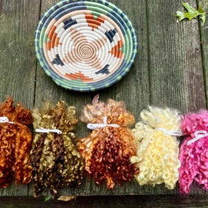 May include: A woven basket with a spiral design in shades of beige, orange, and black sits above six bundles of colorful, curly wool. The wool bundles are tied with white ribbon and come in shades of orange, brown, yellow, and pink.