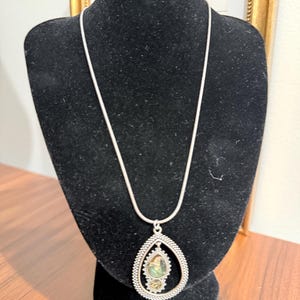 May include: A silver-toned necklace featuring a long, smooth chain and a large, teardrop-shaped pendant. The pendant has a textured, beaded border surrounding a smaller, central teardrop element that contains a mottled, iridescent green stone.
