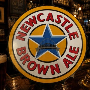 May include: A circular Newcastle Brown Ale beer tap handle featuring a yellow background with a blue star and a grey bridge illustration in the centre. The text NEWCASTLE BROWN ALE is printed in red capital letters around the star design.