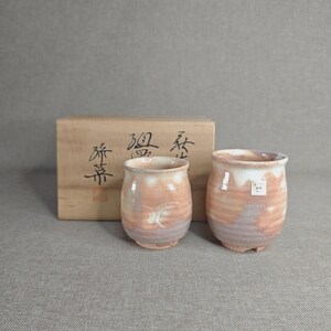 May include: Two ceramic tea cups with a rounded shape and a mottled pink, white, and grey glaze. The cups are positioned in front of a rectangular wooden storage box featuring black Japanese calligraphy characters and a small red seal stamp.