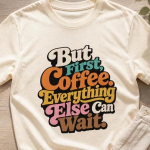 May include: A cream-colored short-sleeve t-shirt featuring a multi-colored graphic print with the text "But First, Coffee, Everything Else Can Wait." The lettering uses a bold, rounded font in shades of black, orange, yellow, pink, and teal.