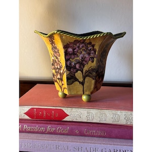 May include: A yellow, flared, hexagonal planter with a scalloped, rope-textured rim and three small, round, green feet. The exterior features a dark purple floral print of hydrangea blossoms and tall, slender, vertical flower stalks.