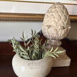 May include: A round, off-white ceramic bowl with a speckled, weathered texture containing a cluster of green, pointed leaves. Beside it stands a tall, decorative stone-colored pinecone sculpture featuring a distressed finish on a square base.
