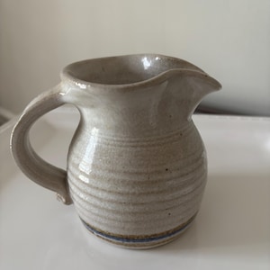 May include: A small, light beige ceramic creamer pitcher with a rounded body, a curved handle, and a pouring spout. The exterior features horizontal ribbed textures and a thin blue band near the base, with a speckled, matte finish throughout.