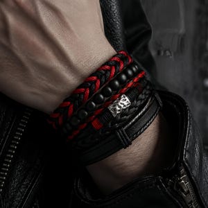 Op de afbeelding: Een meerlaagse armbandset bestaande uit een gevlochten rood en zwart koord, een streng matzwarte kralen en een zwarte leren band met een getextureerd zilverkleurig metalen accentstuk. De set wordt als een gestapeld geheel om de pols gedragen.