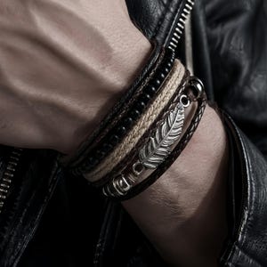 Könnte beinhalten: Ein mehrsträngiges Armband mit einem silberfarbenen Federanhänger aus Metall, einer Reihe kleiner schwarzer Perlen, einer geflochtenen beigen Kordel und dunkelbraunen Lederriemen. Das Design umfasst metallische Akzente und einen Verschluss.