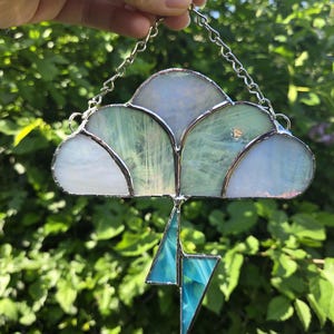 May include: A stained glass suncatcher shaped like a cloud with a hanging lightning bolt. The cloud is made of iridescent white and pale green glass segments joined by silver metal solder. A bright blue glass lightning bolt hangs below.