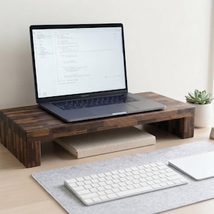 Op de afbeelding: Een donkerbruine houten laptopstandaard met een blokpatroon, waarop een zilveren laptop staat. Onder de standaard ligt een beige notitieboek. Op het bureau staan een wit draadloos toetsenbord, een wit trackpad, een kleine vetplant en een pen in een houder.