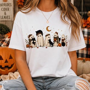 Peut inclure: Un t-shirt blanc à manches courtes orné d'une illustration graphique représentant cinq fantômes et plusieurs chats noirs. Les fantômes portent des accessoires automnaux comme des écharpes et des couronnes, entourés de citrouilles et d'un croissant de lune.