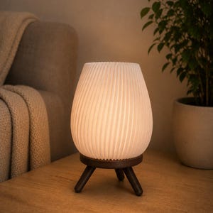 Op de afbeelding: Een tafellamp met een witte, verticaal geribbelde, ronde kap die een warme gloed uitstraalt. De kap rust op een donkerbruine houten voet met drie korte, taps toelopende poten die zorgen voor een stabiele basis voor de verlichting.