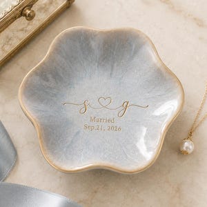 Puede incluir: Un plato decorativo en forma de flor con un acabado brillante marmoleado en azul claro y blanco. El centro presenta texto dorado que dice s corazón g, seguido de la palabra Married y la fecha Sep. 21, 2026, con un borde de color dorado.