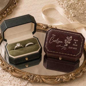 May include: Two octagonal ring boxes, one dark green and one dark red, featuring a velvet interior and gold-tone hardware. The red box lid is engraved with "The Carlson", "Katie & Davis", "22.07.2025", and a floral design. Two rings sit inside.