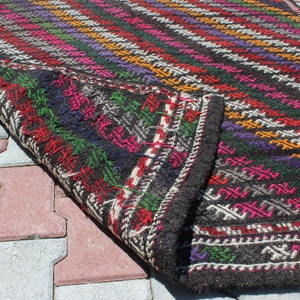 May include: A black wool rug with a colorful geometric pattern. The rug is folded in half and the edges are visible. The rug is lying on a gray and red brick surface.