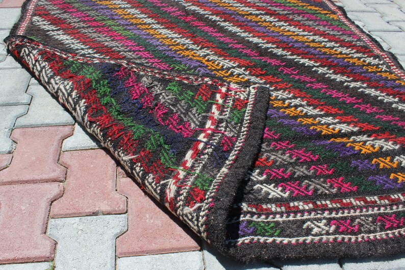 May include: A black wool rug with a colorful geometric pattern. The rug is folded in half and the edges are visible. The rug is lying on a gray and red brick surface.