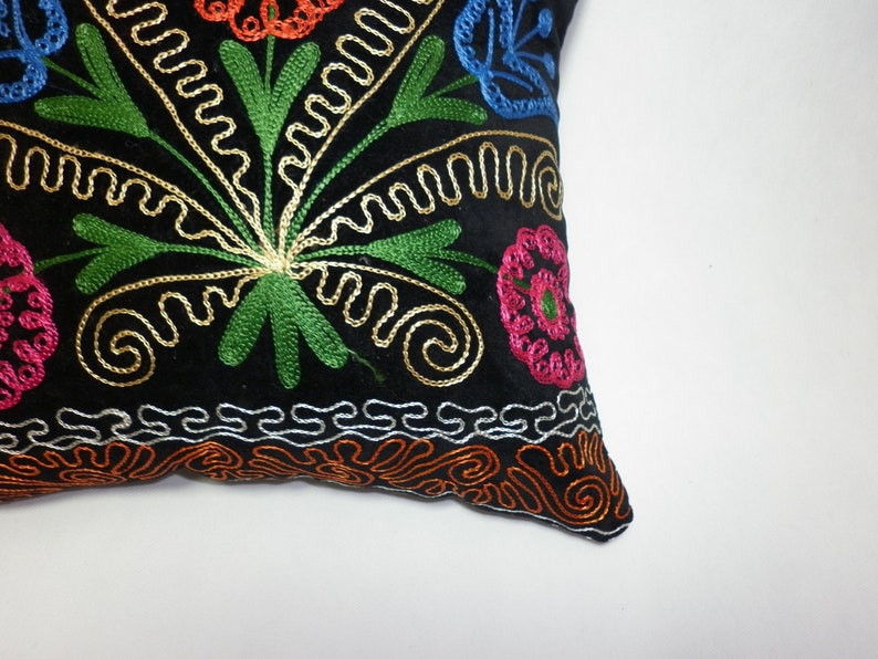 May include: Black velvet pillow with colorful embroidered floral design. The pillow features a central floral motif with green leaves, gold swirls, and pink, blue, and orange flowers.