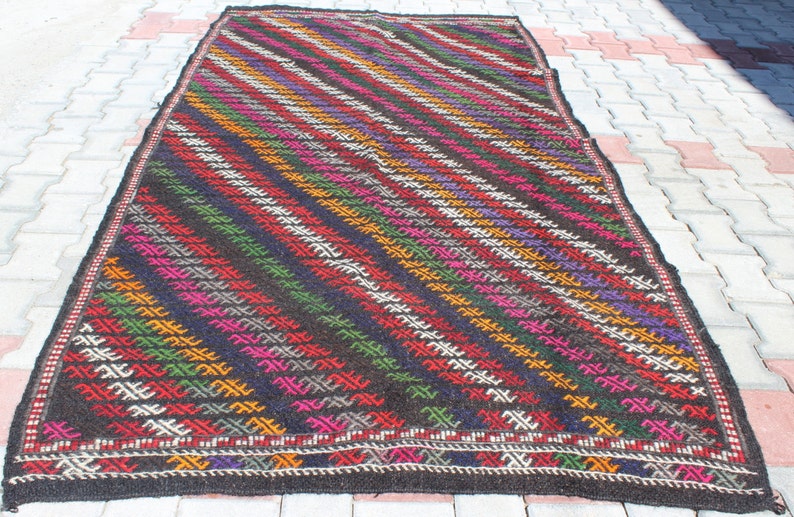 May include: A colorful handwoven rug with a geometric pattern in shades of red, green, yellow, purple, and white on a black background.