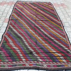 May include: A colorful handwoven rug with a geometric pattern in shades of red, green, purple, orange, and white on a dark brown background.