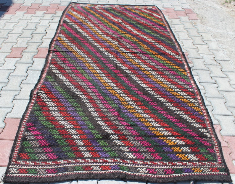 May include: A colorful handwoven rug with a geometric pattern in shades of red, green, purple, orange, and white on a dark brown background.