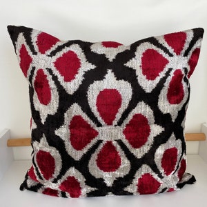 Op de afbeelding: Een decoratief kussen met een zwart, wit en rood ikat-patroon. Het kussen is gemaakt van een zachte, fluweelachtige stof.