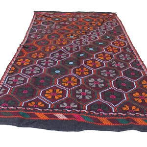 May include: A dark red, black, orange, and green patterned rug with a geometric design of flowers and hexagons. The rug has a black border with a red, orange, and green stripe.