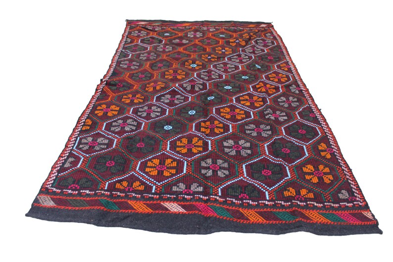May include: A dark red, black, orange, and green patterned rug with a geometric design of flowers and hexagons. The rug has a black border with a red, orange, and green stripe.