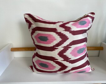 May include: A square pillow with a white background and a geometric pattern in shades of pink, burgundy, and gray.