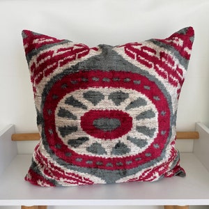 Op de afbeelding: Een decoratief kussen met een rood, wit en grijs ikat-patroon. Het kussen is gemaakt van een zachte, fluweelachtige stof.