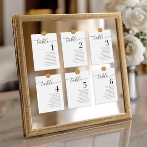 May include: A rectangular mirror with a textured gold frame displaying six white seating chart cards. Each card features the word table followed by a number from one to six and a list of names in black text, secured to the mirror with gold wax seals.