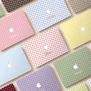 Puede incluir: Una colección de fundas rectangulares para portátiles con varios colores pastel y sólidos con patrones de lunares blancos. Cada funda muestra un logotipo de Apple blanco centrado y un nombre personalizado en letra cursiva, como Catalina, Eleanor y Samuel.