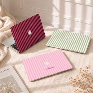 May include: Three laptop skins featuring vertical stripes in burgundy, sage green, and light pink. The pink skin displays the name Jelena in a cursive font. Each skin includes a cutout for the Apple logo, showcasing the metallic silver emblem.