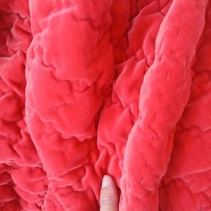 Puede incluir: Una tela de terciopelo acolchada de color rojo vibrante con una textura suave y afelpada, que presenta patrones de costura visibles que crean una serie de secciones acolchadas y elevadas en toda la superficie, resaltando el color intenso.