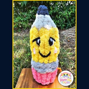 May include: A plush pencil toy made of yellow, gray, pink, and dark blue yarn. The pencil features a smiling face with black embroidered eyes, a mouth, and pink cheeks. The text "Professor Pencil" and "Handknitting Pattern & Tutorial" appear.