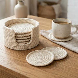 May include: A set of round, cream-colored crocheted coasters stacked inside a matching circular holder. Two additional coasters lie flat on the surface, and one coaster sits beneath a speckled ceramic mug filled with a light brown beverage.