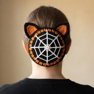 Op de afbeelding: Een ronde gehaakte knot-cover met een wit spinnenwebpatroon op een zwarte achtergrond, omrand door een oranje en zwarte rand, en bekroond met twee puntige kattenoren van oranje en zwart garen, ontworpen om over een haarknot te passen.
