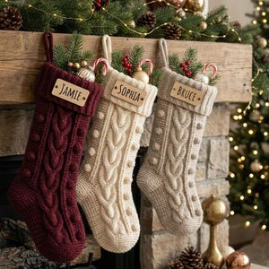 May include: Three knit Christmas stockings in burgundy, cream, and beige, each featuring a cable knit pattern with bobbles and a rectangular wooden name tag labeled JAMIE, SOPHIA, or BRUCE. Each stocking contains pine branches and candy canes.